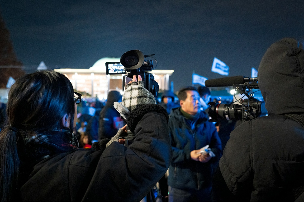 A group of people standing at a political event and two media people with cameras